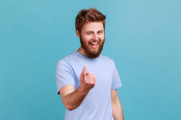 Man in blue T-shirt making beckoning gesture, inviting to come, flirting and looking playful. Come here. Portrait of bearded man making beckoning gesture, inviting to come, flirting and looking playful, having excited facial expression. Indoor studio shot isolated on blue background. beckoning photos stock pictures, royalty-free photos & images