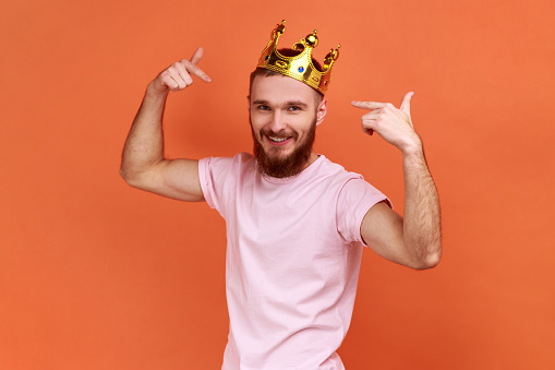 Portrait of smiling handsome bearded man pointing fingers on golden crown on his head, showing his authority, wearing pink T-shirt. Indoor studio shot isolated on orange background.