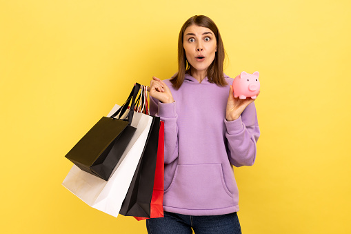 Portrait of astonished woman with open mouth holding shopping bags and piggy bank, save big mount of money, great purchase, wearing purple hoodie. Indoor studio shot isolated on yellow background.