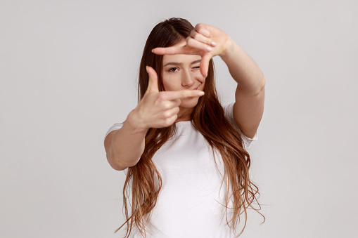 Woman looking through finger frame, imitating to take photo, focusing and cropping interesting moment, capturing moment, wearing white T-shirt. Indoor studio shot isolated on gray background.