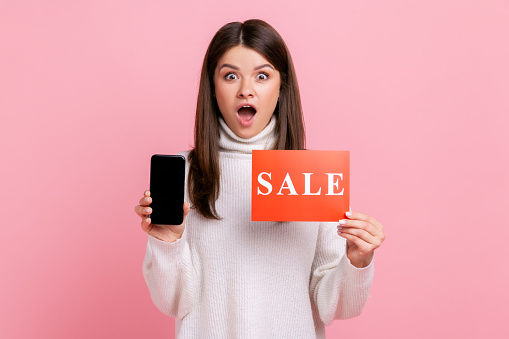 Astonished female standing with open mouth, holding sale card and cell phone with empty display, wearing white casual style sweater. Indoor studio shot isolated on pink background.