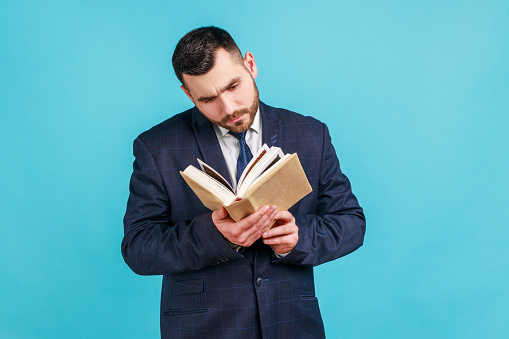 Young adult concentrated man wearing dark suit reading book, enjoying reading, standing absorbed with exciting plot, checking schedule in notepad. Indoor studio shot isolated on blue background.