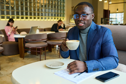 Portrait of African businessman in eyeglasses smiling at camera while sitting at table in cafe and drinking coffee