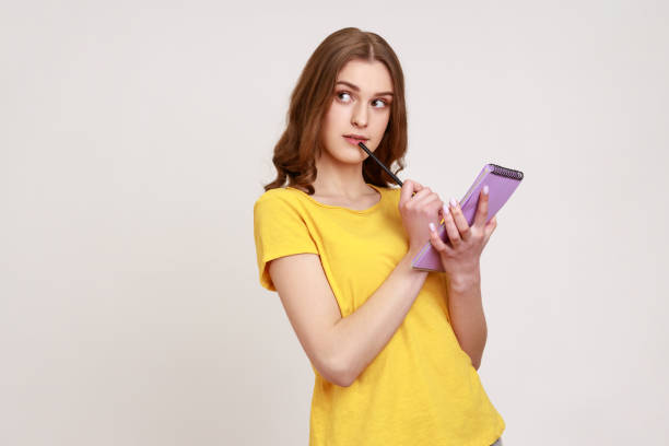 beautiful thoughtful teen girl in yellow casual style t-shirt holding notebook in hand, writing down plans for future, thinks about to do list. - pensive question mark teenager adversity imagens e fotografias de stock