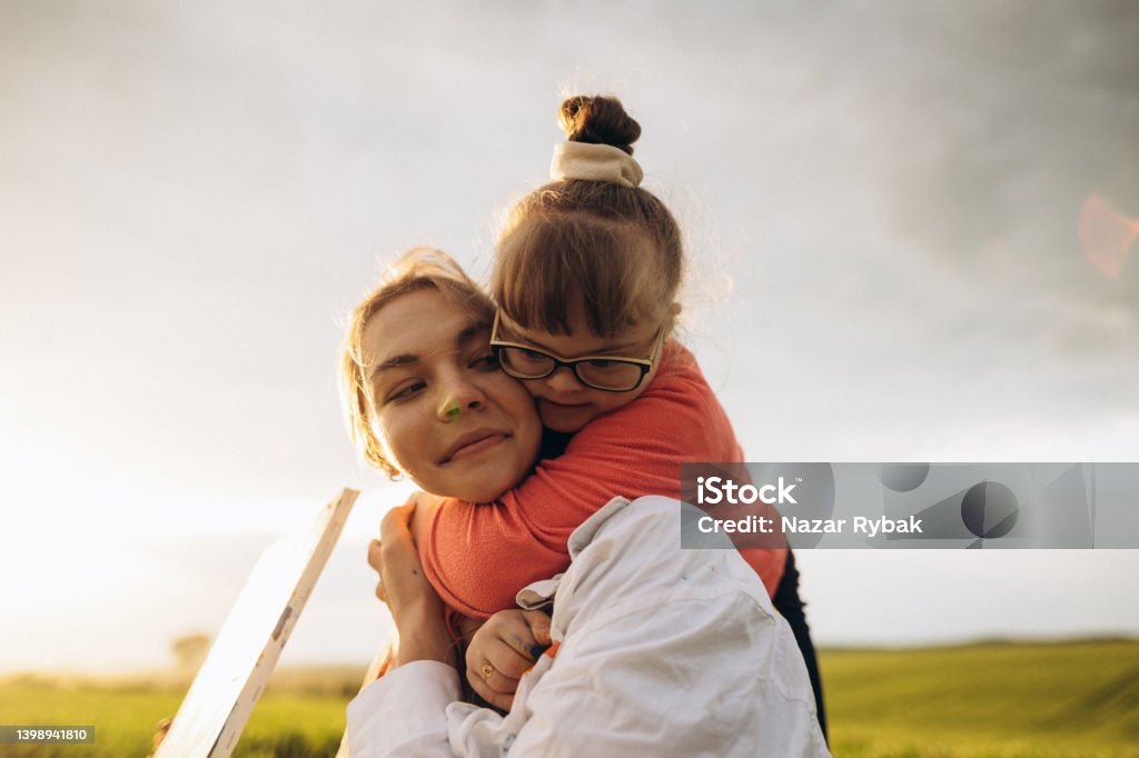 A Downs syndrome girl hugs a woman A Downs syndrome girl hugs her female art therapist at the sunset Down Syndrome Stock Photo