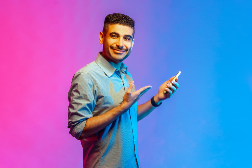 Side view of happy optimistic man in shirt using cell phone, smiling, showing his mobile phone, enjoying mobile app. Indoor studio shot isolated on colorful neon light background.