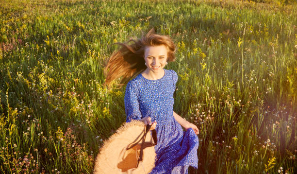 Artistic image of an adult woman sitting in the grass in a long dress and hat on her head.