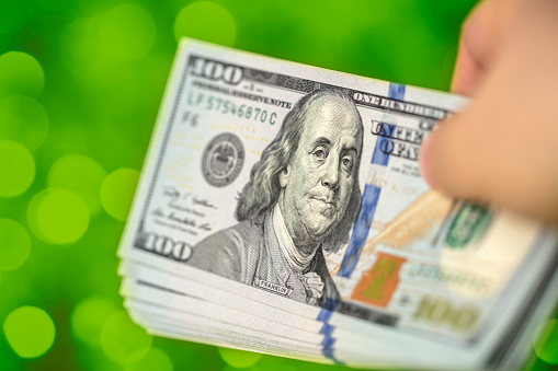 Close-up of a hand holding a bundle of US $100 bills. Green background with beautiful defocused lights bokeh. Shallow depth of field.