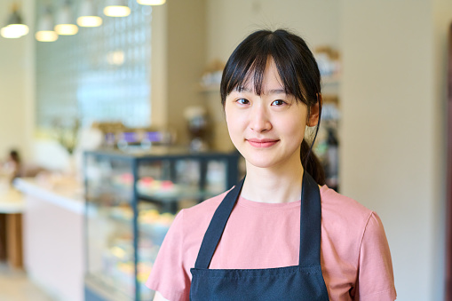 Portrait of young Asian waitress in uniform looking at camera standing in coffee shop