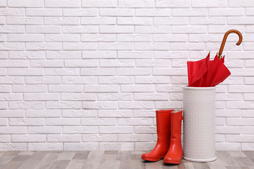 Red umbrella in holder and rubber boots near white brick wall indoors. Space for text