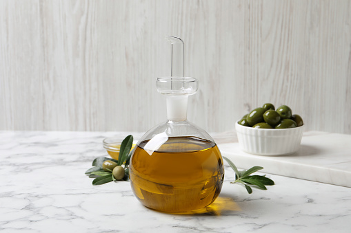 Glass jug of oil, ripe olives and green leaves on white marble table