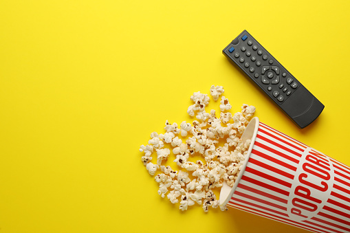 Remote control and cup of popcorn on yellow background, flat lay. Space for text