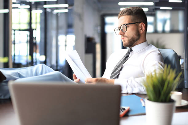 istock Handsome businessman sitting with legs on table and examing documents in office 1398960097