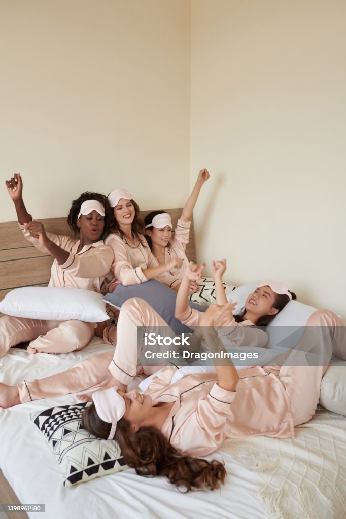 Female Friends Kidding Around Diverse group of female friends in satin pajamas kidding around on bed Sorority Stock Photo