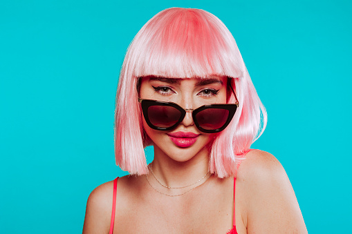 Young attractive female in pink wig and sunglasses, posing at studio over blue background. Face shot portrait. Glamour, fashion concept.