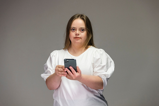 Young adult female with down syndrome holding her mobile phone with a blank expression looking at camera standing in front of a grey background.