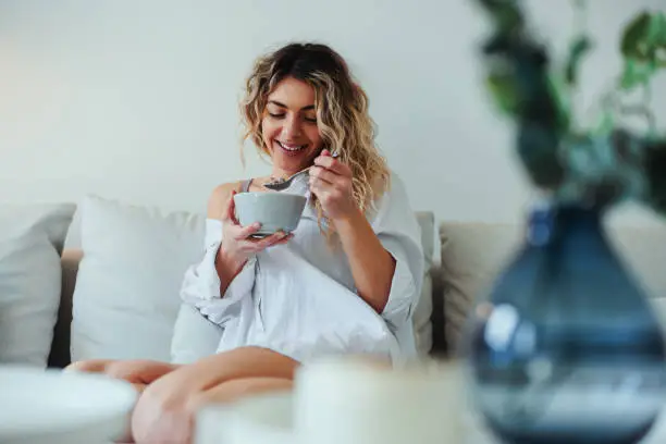 Smiling woman sitting on sofa and eating healthy breakfast at home in morning