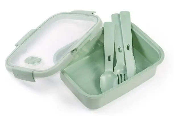 Empty reusable food storage container with partly removed transparent lid and plastic spoon, fork and knife inside on a white background
