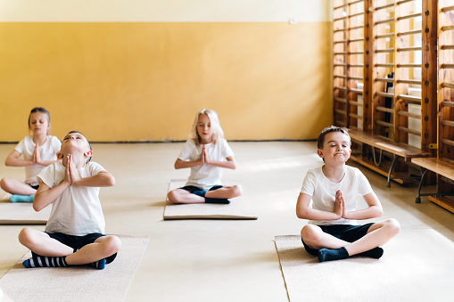 elementary school children sitting cross-legged on the floor of gym and meditating in yoga lesson