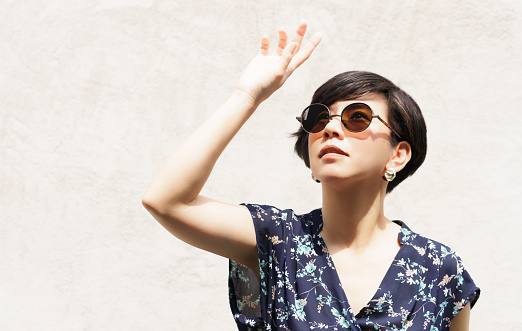Stylish short hair Asian woman with sunglasses raising hand to block sunlight with clean cement wall in background in sunny day