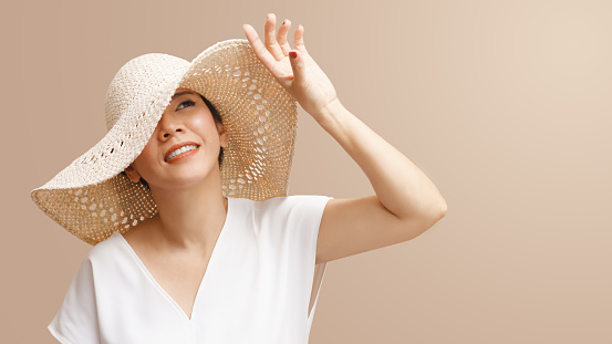 Beautiful short hair Asian woman with white top, straw hat looking up, smiling and raising hand to block sunlight