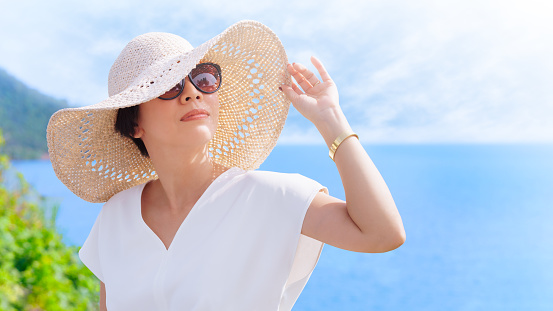 Beautiful short hair Asian woman with white top, straw hat and sunglasses looking up, smiling and raising hand to block sunlight at the beach with blue sea and sky.