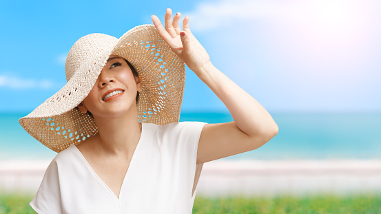 Beautiful short hair Asian woman with white top, straw hat looking up, smiling and raising hand to block sunlight at the beach with blue sea and sky.