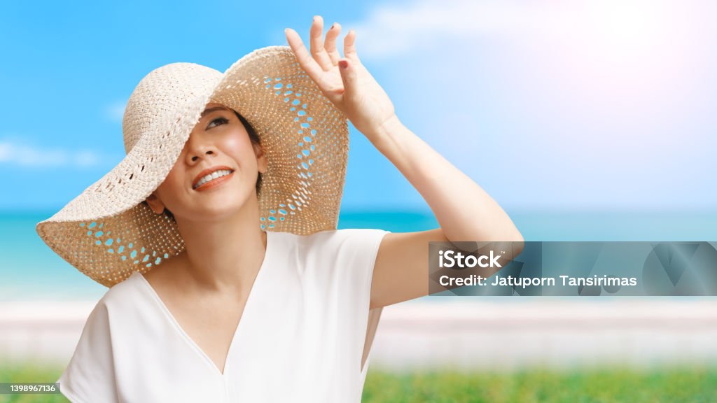Beautiful Asian woman smiling and raising hand to block sunlight at the beach Beautiful short hair Asian woman with white top, straw hat looking up, smiling and raising hand to block sunlight at the beach with blue sea and sky. Sunburned Stock Photo