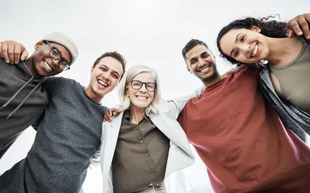 Photo of Low angle portrait of team of business people hugging each other. Diverse group of men and women standing in a together in their office, huddling and smiling. Unity and teamwork is key to success