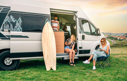 Group of friends traveling in a camper van. Two women sitting in front of camper van while young man cooks