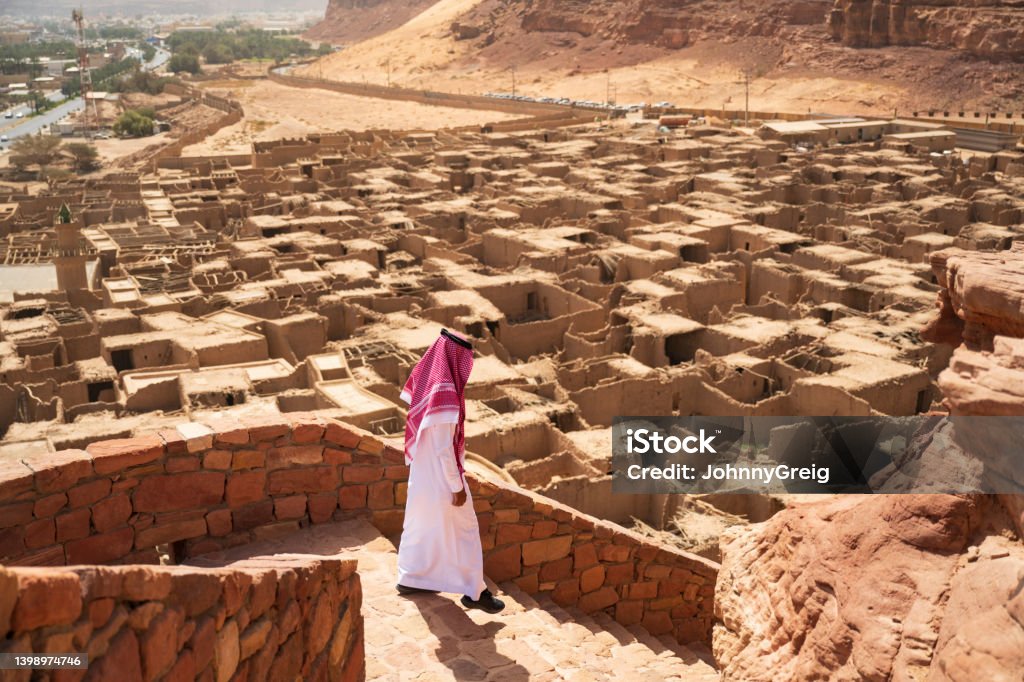 Elevated viewpoint across Al-Ula Old Town Full length view of man in traditional attire moving down steps of ancient Saudi Arabian fort with mudbrick houses and desert oasis in background. Saudi Arabia Stock Photo