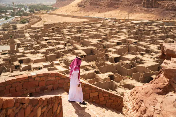 Full length view of man in traditional attire moving down steps of ancient Saudi Arabian fort with mudbrick houses and desert oasis in background.