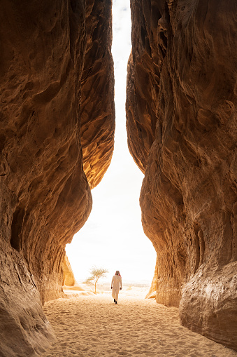 Full length view of smiling Middle Eastern man in traditional attire approaching camera within Jabal Ithlib’s natural narrow passageway.