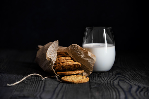 A glass of milk and muesli biscuits in a paper bag on a dark background. The wood texture of the countertop is visible. Concept. Background. Rustic. Dinner.