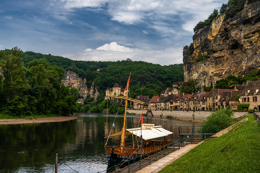 La Roque-Gageac, France - 12 May, 2022: view of the historic village of La Roque-Gageac with the Dordogne River and cliffside houses and a wooden Gabare river boat in the foreground