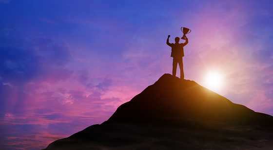 Silhouette victory businessman with trophy climbed to the top of mountain over the sky and sun light background. Success, Achieving goal, Leadership, Career, Winner, and Business achievement Concept.