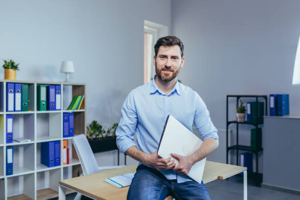 Portrait of a young teacher in a classroom, a man sitting at a desk, holding a laptop, looking at the camera and smiling Portrait of a young teacher in a classroom, a man sitting at a desk, holding a laptop, looking at the camera and smiling professor business classroom computer stock pictures, royalty-free photos & images