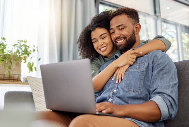 giovane coppia di razza mista felice sorridente mentre usa un laptop insieme a casa. gioioso fidanzato ispanico e fidanzata che si rilassano e usano un laptop nel salone di casa - hometeam foto e immagini stock