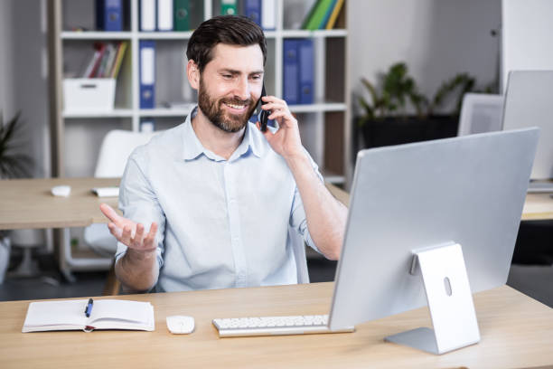 istock Young man with a beard, freelancer, manager, worker talking on a cell phone in the office, sitting at a computer desk, talking, waving 1398991102