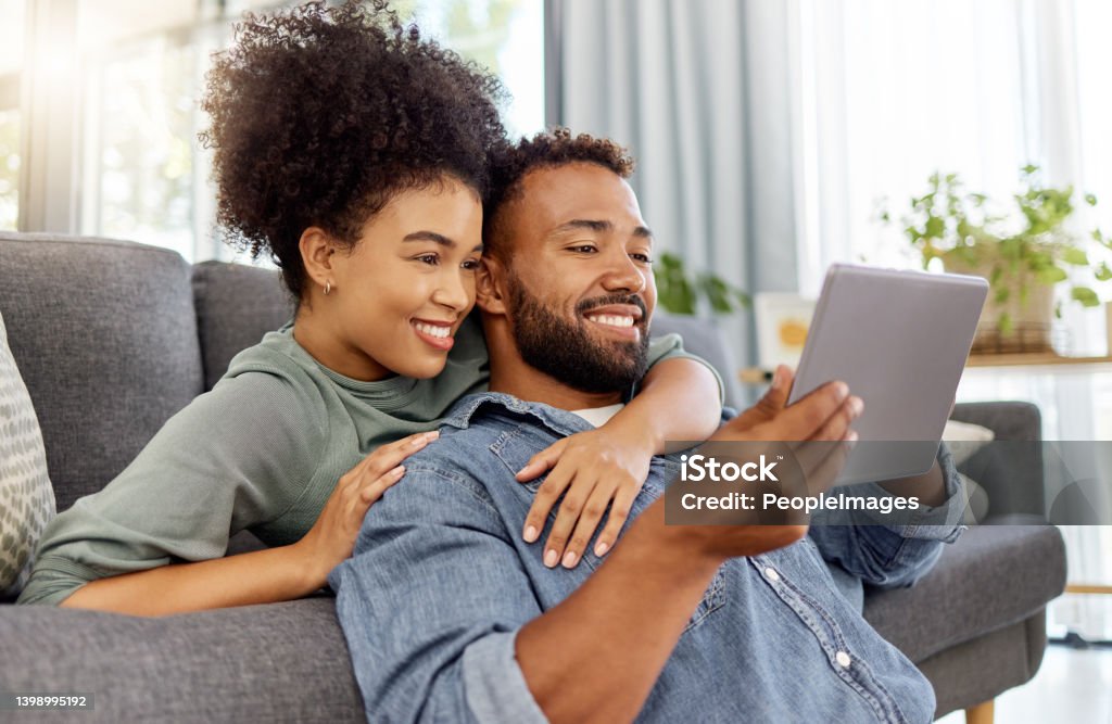 Mixed race couple smiling while using a digital tablet together at home. Content hispanic boyfriend and girlfriend relaxing and using social media on a digital tablet in the lounge at home Couple - Relationship Stock Photo