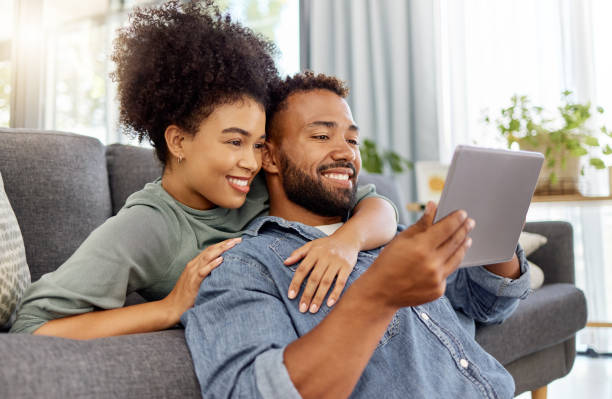 mixed race couple smiling while using a digital tablet together at home. content hispanic boyfriend and girlfriend relaxing and using social media on a digital tablet in the lounge at home - olhando imagens e fotografias de stock