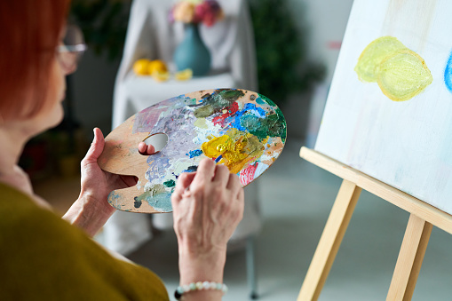 Close-up of woman mixing paints with paintbrush on palette and painting picture on canvas during lesson