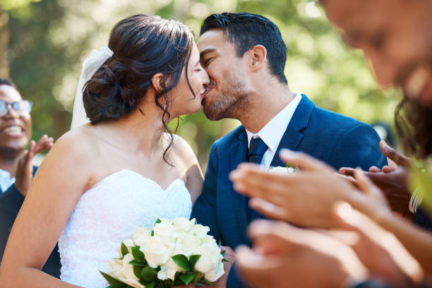 felice coppia romantica di sposi che si bacia fuori mentre è circondata da amici e familiari nel giorno del loro matrimonio. sposa e sposo profondamente innamorati - real estate immagine foto e immagini stock