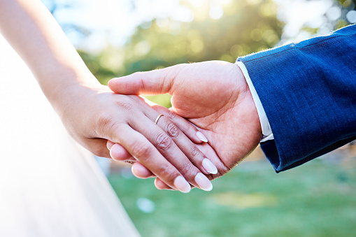 Close up hands of newlywed couple standing outside on a sunny day. Groom holding his brides hand with wedding band
