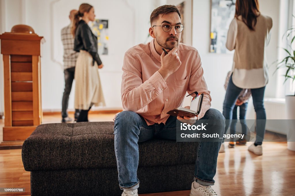 Man enjoying art Diverse group of visitors in modern art gallery, man sitting, taking notes and looking at paintings. Critic Stock Photo