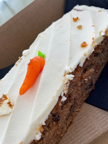 Stock photo showing close-up view of single-use cardboard container with carrot cake slice on black paper serviette. The carrot cake is topped with a rich cream cheese icing and decorated with an orange fondant carrot decoration.