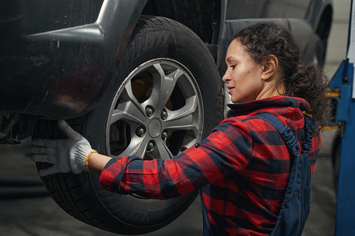 Female vehicle technician in work gloves changing automobile wheel in repair service station