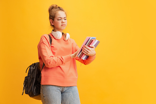 Young girl, student with dissatisfied facial expression looking on tablet, posing with school supplies isolated over yellow background. Concept of education, studying, homework, youth, lifestyle