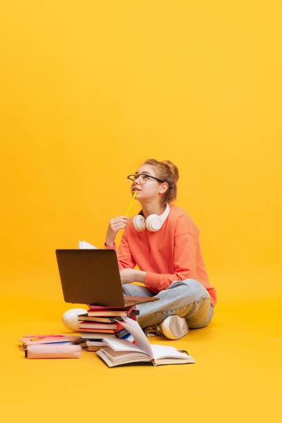 ritratto di giovane ragazza, studentessa in occhiali seduta sul pavimento, che studia isolata su sfondo giallo - homework teenager education mobile phone foto e immagini stock