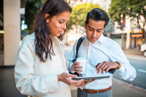 Hispanic young adult coworkers drinking coffee and discussing business ideas before work. Carrying a digital tablet and a smartphone while standing on the streets of Los Angeles.